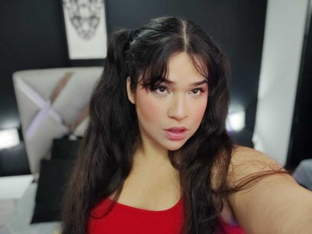 AntonelaPark with long dark hair in pigtails, red dress, pink eye makeup, and pink lipstick, looking at camera, serious expression, visible shoulder, black and white background.