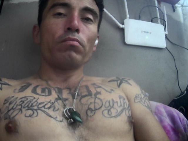 BootyHotMan is a shirtless dude with tattoos on his chest, wearing white earbuds, and a green pendant. He has a serious face and short black hair.