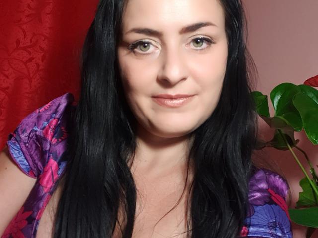 MistressMayaDiamondXx has pale skin, green eyes, black hair, and is wearing a purple floral top. Her small tits are slightly visible. Red curtain and green plant in background.