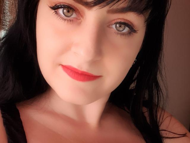 MistressMayaDiamondXx with pale skin, black hair, red lipstick, and brown eyes, smiling at the camera, wearing a black top, showcasing her tits.