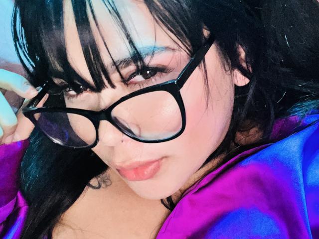 Melina69, avec ses lunettes noires, ses cheveux noirs lisses, sa frange et son rouge à lèvres rose, porte une chemise violette, a un petit anneau au nez et un tatouage sur son épaule droite..