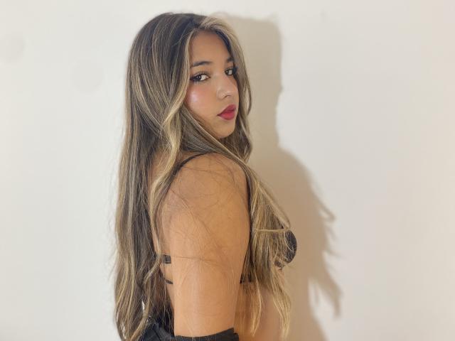 AmbarDunn with long blonde and brown ombre hair, red lipstick, black bra, and side glance, showing off her tits against a white wall.