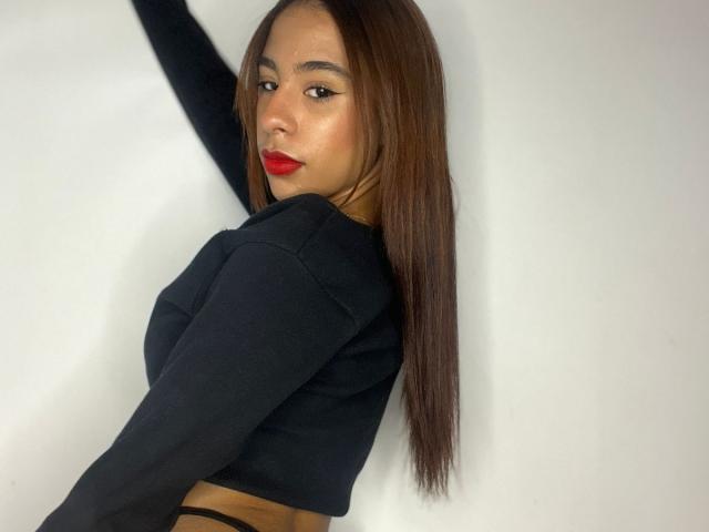 LisshaCute with long brown hair, wearing a black long-sleeve top, showing her back and looking over her shoulder, red lips, and a slight smile, exposing her black strappy bottom.