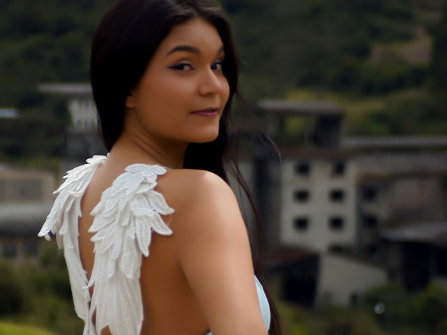AriaLeenn with dark brown hair, tan skin, and white feathered dress, showing her back and looking over her shoulder with a slight smile. Boobs visible in light blue top.