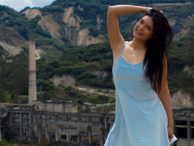 AriaLeenn, smiling, wearing a light blue, semi-transparent dress, has her hand in her long black hair, showing her tits, standing in front of a ruined building and green hills.
