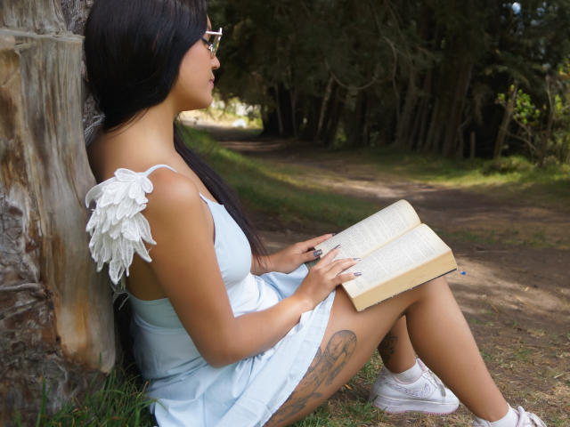 AriaLeenn with dark hair, glasses, white wings, light blue dress, tattoo on thigh, sitting against tree, reading book, holding pen, wearing white sneakers, outdoors, daytime.