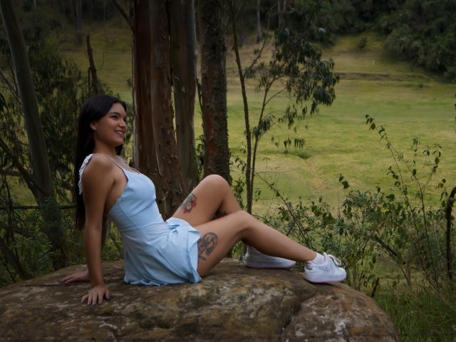 AriaLeenn with medium tits, sitting on a rock, wearing a light blue dress, white sneakers, showing tattoos on her thighs, smiling, with dark hair, in a forest.
