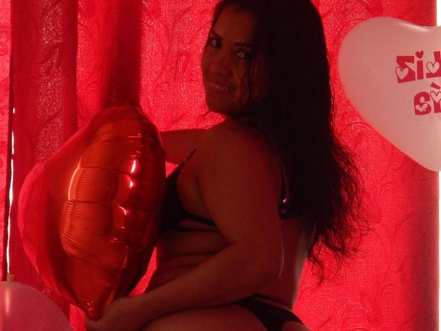 NatashaMonttero with long dark hair, black lingerie, holding a red balloon, smiling, red curtain, white balloon with red symbols, dim red lighting, sexy, curvy, attractive, looking over shoulder.