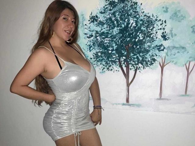 HotPamelaSex with large tits in a shiny, silver, tight dress, hands on hips, smiling, long brown hair, standing, painting of trees in background.