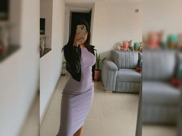 AnyaJay with black leather jacket, purple dress hugging her tits and ass, standing in front of mirror, holding phone with colorful case, in modern living room.