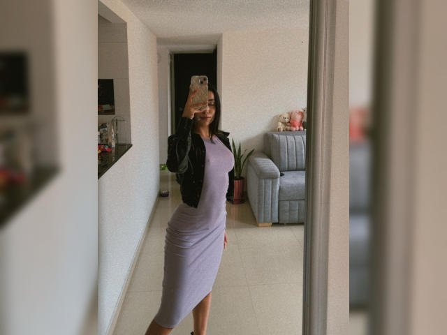 AnyaJay with curvy body, small tits, and a purple dress, taking a mirror selfie, smiling, wearing a black jacket, in a modern living room with a gray sofa.