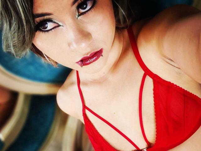 CassieJones with red lingerie, looking up with seductive eyes, full red lips, and pierced lower lip. Her tits are small, and she has a slim, attractive build.