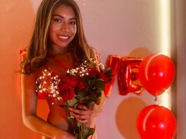 EimyRousse mit langen, braunen Haaren, die rote Rosen und Babysbreath hält, trägt rote Dessous, lächelt und ist umgeben von roten Ballons und glänzenden "LIEBE"-Lettern..