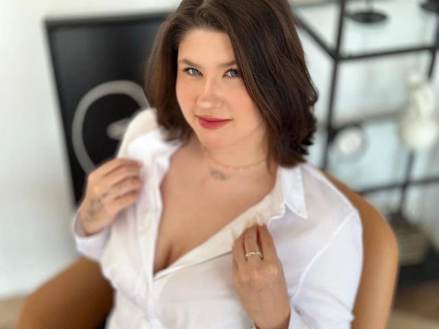 AmyAmethyst-hot with brown hair, white shirt, cleavage, small tattoos on hands, subtle smile, ring on right hand, seated, looking at camera, slightly open shirt.
