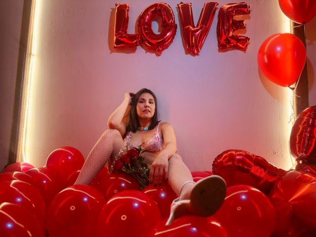 AdelaShery with her tits in a pink bra, sitting on red balloons, wearing fishnet stockings, holding roses, surrounded by red balloons and shiny heart-shaped decorations, looking sexy.