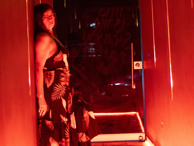 AntonellaDupontt está de pie en un pasillo iluminado con luz roja, apoyada contra una pared, vestida con un vestido negro con estampados florales, con las manos apoyadas en sus glúteos, mirando por encima del hombro..