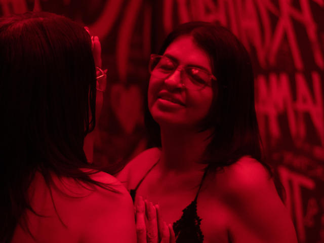 AntonellaDupontt with red glasses, smiling at another person with long dark hair, both in black lace lingerie, hands touching, red lighting, intimate, sensual.