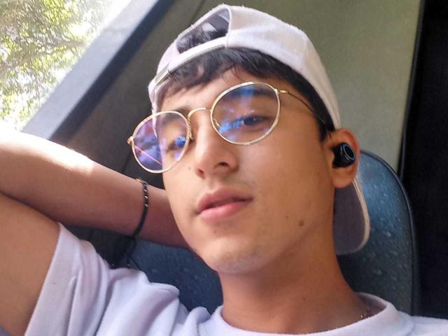 DeyerHot69, usando óculos de aviador dourados com efeito reflexivo, um boné branco, fones de ouvido pretos e com uma expressão relaxada, com os braços atrás da cabeça, vestindo uma camiseta branca, dentro de um carro..