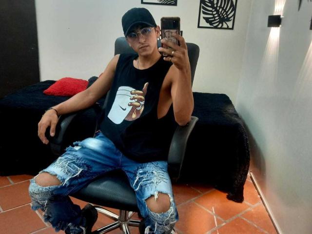 DeyerHot69, que lleva una camiseta negra con un estampado de un teléfono blanco, jeans azules rotos, una gorra negra y gafas, está sentada en una silla, tomando una foto de sí misma en el espejo..