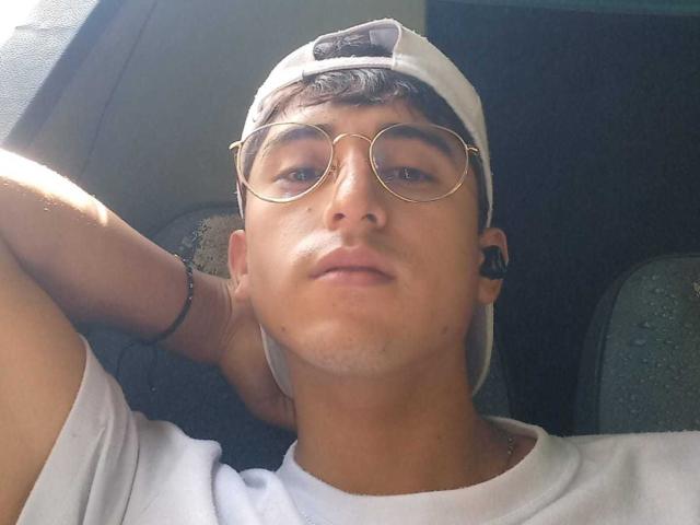 DeyerHot69, com uma expressão relaxada, usando um boné branco, óculos redondos, camisa branca e fones de ouvido pretos, apoiado no assento do carro, iluminado pelo sol, com um leve sorriso..