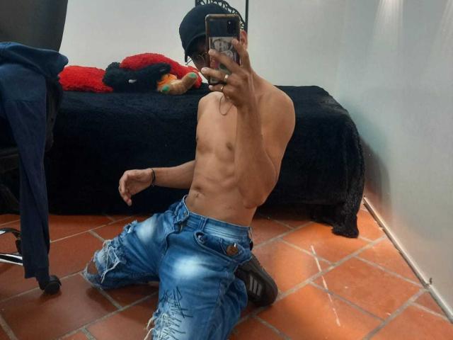 DeyerHot69 is shirtless, kneeling on orange tile, wearing blue ripped jeans, black cap, holding phone, showing a slim, toned torso with a visible ribcage.