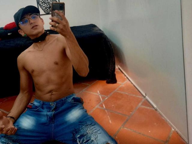 Shirtless DeyerHot69 with a slim, toned body, wearing a black cap, glasses, and a choker, sitting on orange tile floor, taking a mirror selfie, in blue ripped jeans.