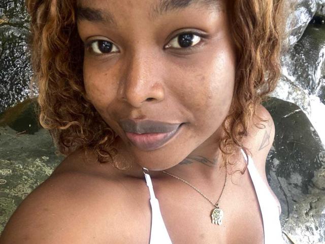 MadinsonLopez has dark brown skin, curly blonde hair, and is wearing a white tank top, showing her small tits and grey lipstick. She's looking at the camera with a smile.