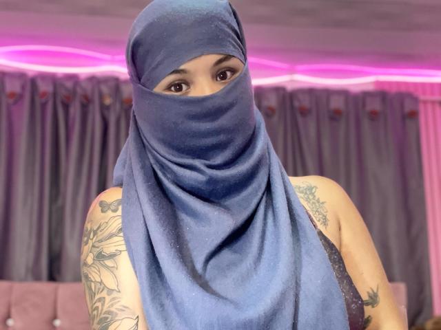 MalakCote with a blue headscarf covering her face, showing brown eyes, tattoos on her arms, and wearing a sleeveless top in a pink-lit room.