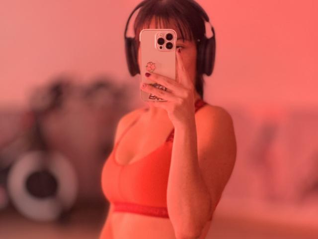 Liliysweet with red bra, holding phone to her face, wearing headphones, blurred background, red lighting, medium breasts, fair skin, dark hair, phone case with small red sticker.
