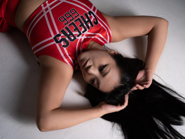 DragonDiva is lying on her back with black hair spread out, wearing a red cheerleader top, arms above her head, looking up with a serious expression.