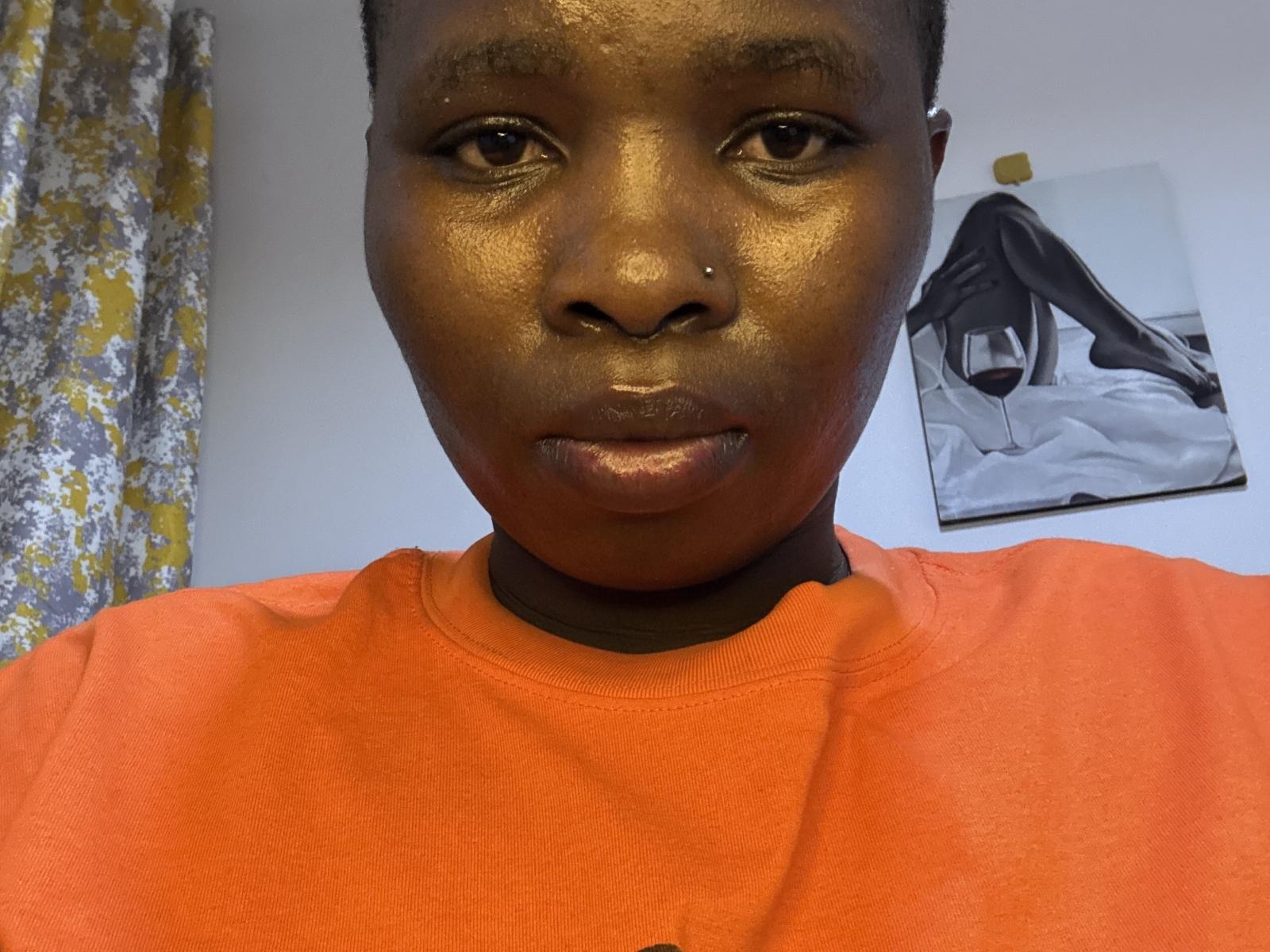 KenyanHotGirl with shiny dark skin, short curly hair, pierced nose, and a red shirt, showing her shiny lips and glistening face. Background includes a yellow and gray curtain.