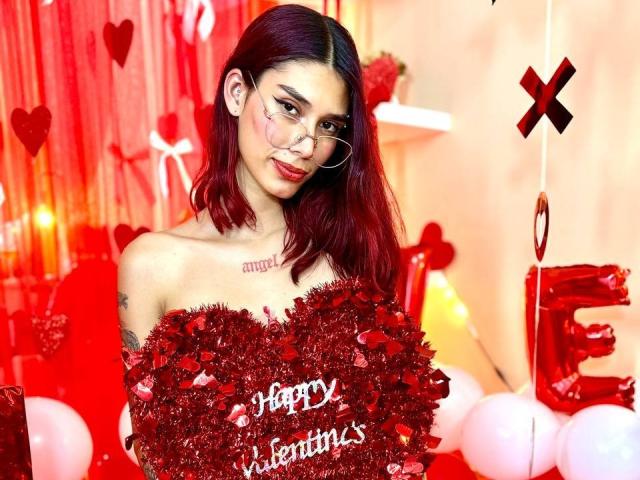 NycoleAngels with red hair, tattoos, and glasses, holding a glittery red heart with "Happy Valentine's" in front of her, surrounded by red balloons and heart decorations.
