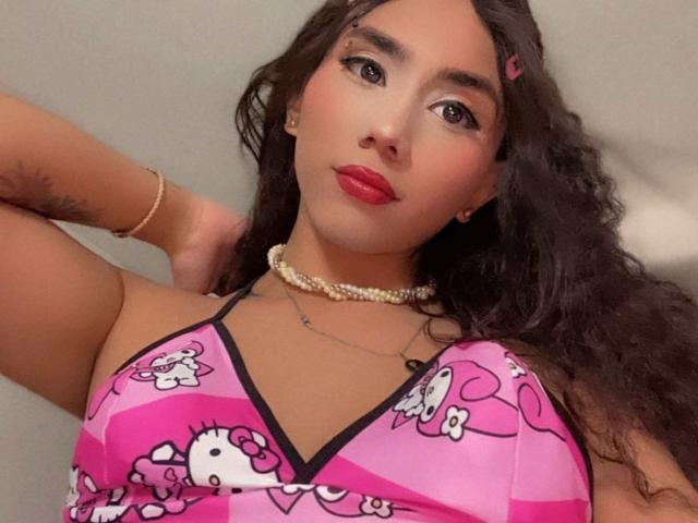 MiaSoldatty, com cabelo preto encaracolado, batom vermelho e um vestido rosa do Hello Kitty, usando um colar e pulseira de pérolas, mostrando uma tatuagem no braço. Seios visíveis..