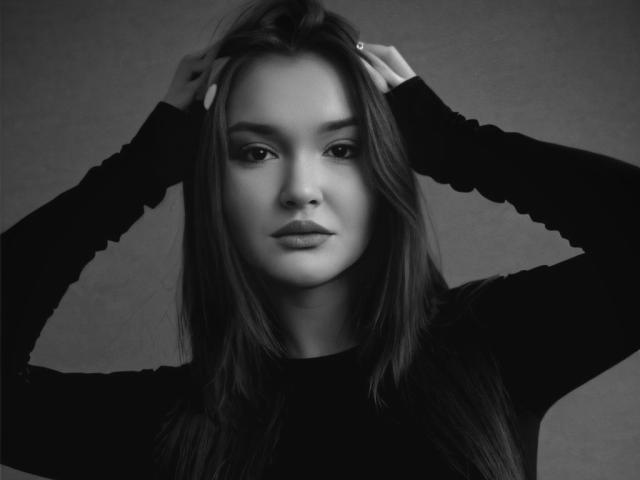 EvaLacroix with hands in long, straight brown hair, wearing a black long-sleeve shirt, looking directly at the camera, black-and-white, serious expression.