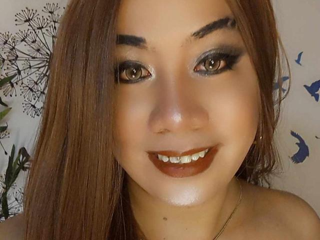 TSGorgeousHotCum has brown eyes, dark smoky eye makeup, shiny skin, brown lipstick, straight brown hair, and a cute smile with visible teeth. Her ass is not visible.