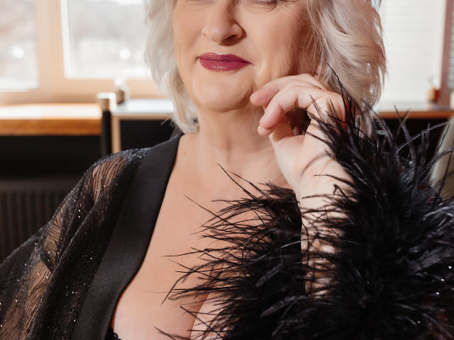LolkaAlyss has white curly hair, purple lipstick, and a black sheer dress with sparkles. She's holding a black feather, showing her tits slightly. Her finger touches her cheek.