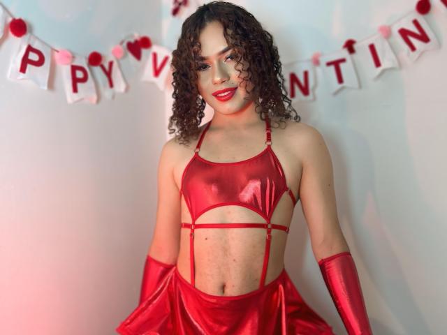 SexyMonroe with curly brown hair, wearing a red metallic halter top and matching gloves, showing a flat stomach with a cut-out, and a red skirt, smiling with red lipstick.