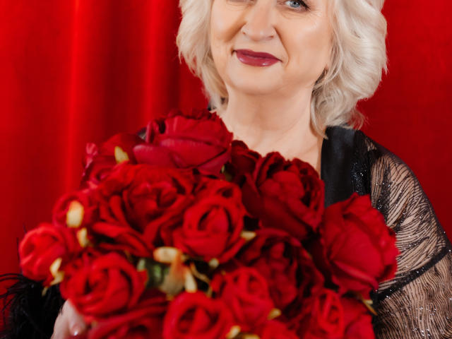 LolkaAlyss, avec ses cheveux blancs bouclés, son rouge à lèvres rouge et sa robe en dentelle noire, tenant un bouquet de roses rouges, souriant à l'appareil photo..