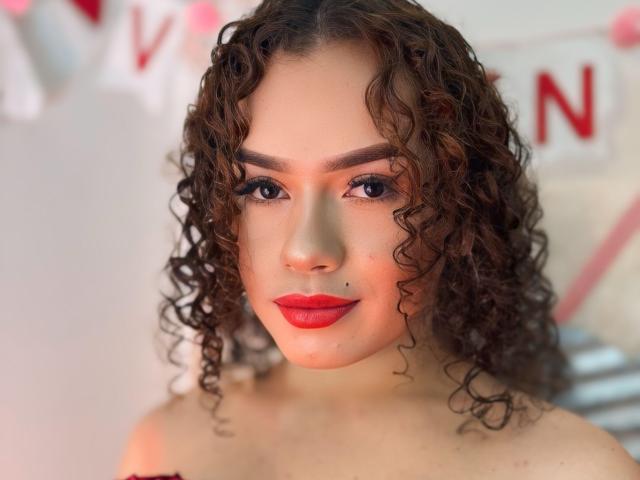 SexyMonroe with curly brown hair, red lipstick, and a small mole on her upper lip, staring confidently at the camera, topless, showing bare shoulders, in a well-lit photo.