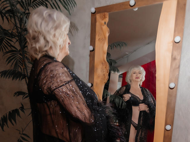 LolkaAlyss with white curly hair, black sheer lingerie, and black feather robe, stands in front of a mirror, revealing her tits and black bra in a mirror selfie.