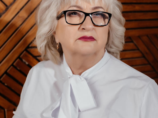 LolkaAlyss tem cabelo branco, óculos pretos e veste uma camisa branca com um laço. Ela tem batom rosa e uma expressão séria. Seus seios são pequenos e ela é velha..
