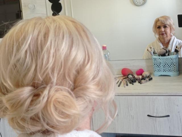 Blond BozhenaNude with curly updo stands in front of mirror, reflection shows her smiling face, white blouse, blue patterned holder with makeup tools, pink and red balls on counter.