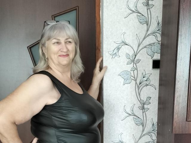 LolkaAlyss, una donna di mezza età con i capelli grigi, indossa una maglia nera dall'aspetto in pelle, è in piedi in una porta, con una mano appoggiata al muro, carta da parati con motivi floreali, sorride, mostrando il suo seno prosperoso..