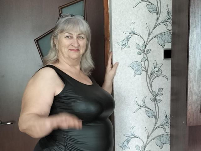 LolkaAlyss, middle-aged woman with gray hair, wearing a black leather dress, standing in a doorway, touching floral-patterned wall, slightly smiling, showing her tits and ass.