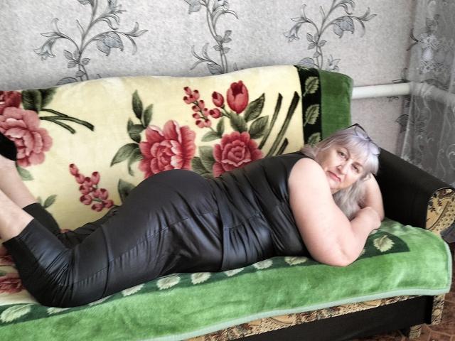 LolkaAlyss, lying on a green and floral-patterned sofa, wearing a black leather dress, showing her ass, with white hair, looking at the camera, legs up, arms under chin.