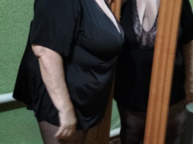LolkaAlyss with large boobs in a black dress and lace bra, wearing black stockings, standing in front of a mirror. Her reflection shows her cleavage.
