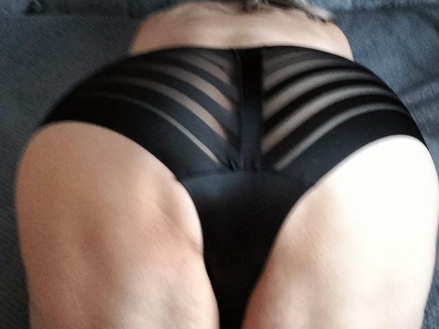 LolkaAlyss ligt op haar buik, gekleed in zwarte, doorschijnende onderbroeken met strepen, waardoor haar ronde billen zichtbaar zijn. Haar huid is bleek, en de aandacht is gericht op haar achterwerk..
