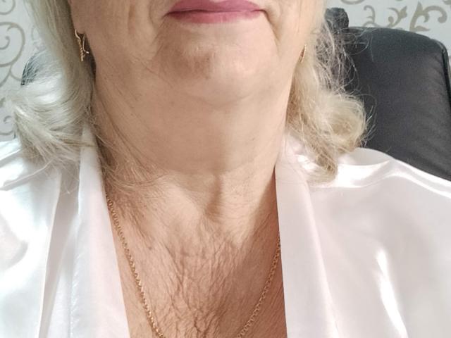 Le visage de LolkaAlyss, celui d'une vieille dame, montre un cou ridé, du rouge à lèvres rose, une robe en satin blanc, des boucles d'oreilles en or et un collier en or. Ses cheveux gris sont ondulés, et elle regarde vers le bas..