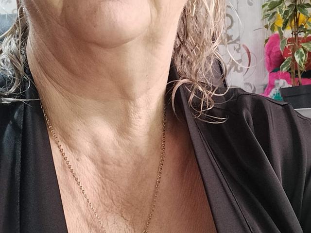 Le cou et la partie supérieure de la poitrine de LolkaAlyss, ornés d'un collier en chaîne dorée, présentent un pli profond et quelques rides. Elle porte un haut noir, légèrement ouvert, en soie..