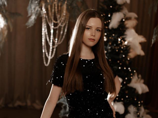 MileyGloss with long brown hair, wearing a black sequined dress, standing in front of a decorated Christmas tree, looking confident with a slight smile, medium-sized tits, cute face.